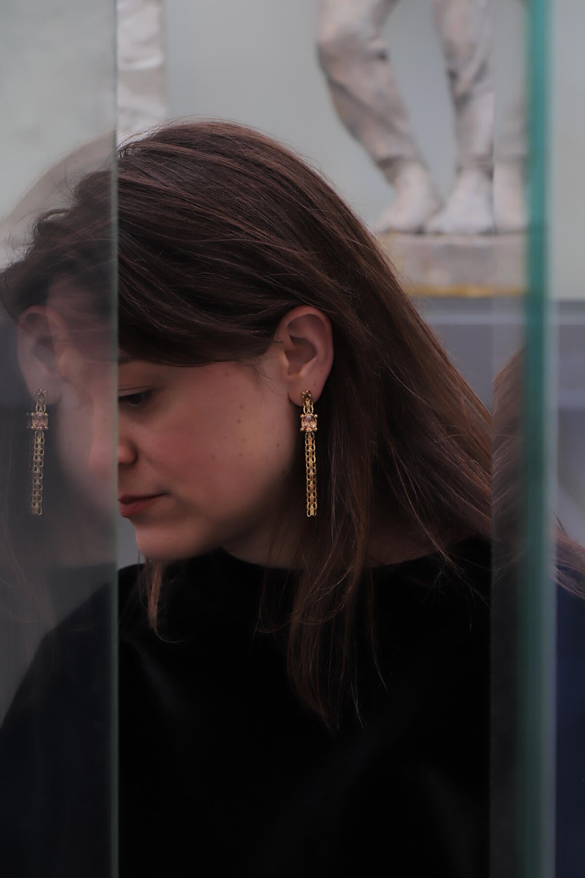 Saskia wearing Drip earrings with oval-cut orange cubic zirconia reflected in a glass display at La Piscine Musée Roubaix.