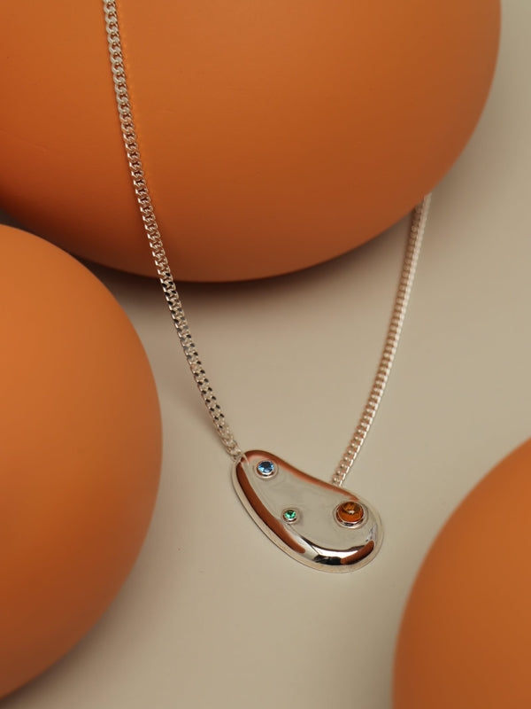 Spill necklace with cubic zirconia featuring a sculptural pendant set with blue, green and orange stones on a smooth orange setting.