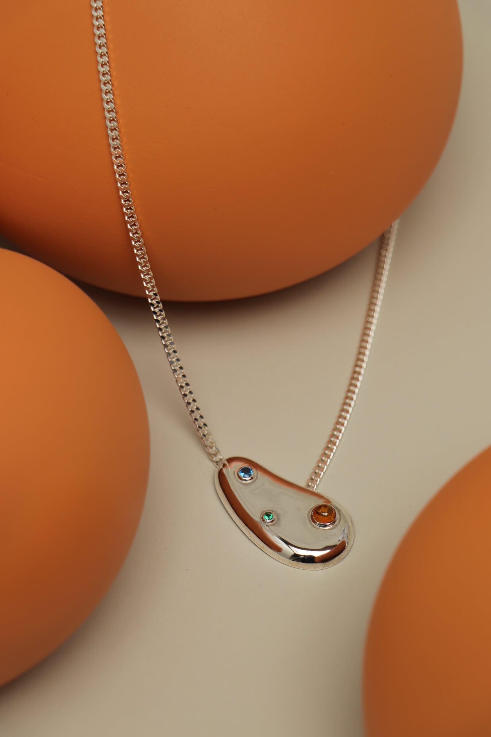 Spill necklace with cubic zirconia featuring a sculptural pendant set with blue, green and orange stones on a smooth orange setting.