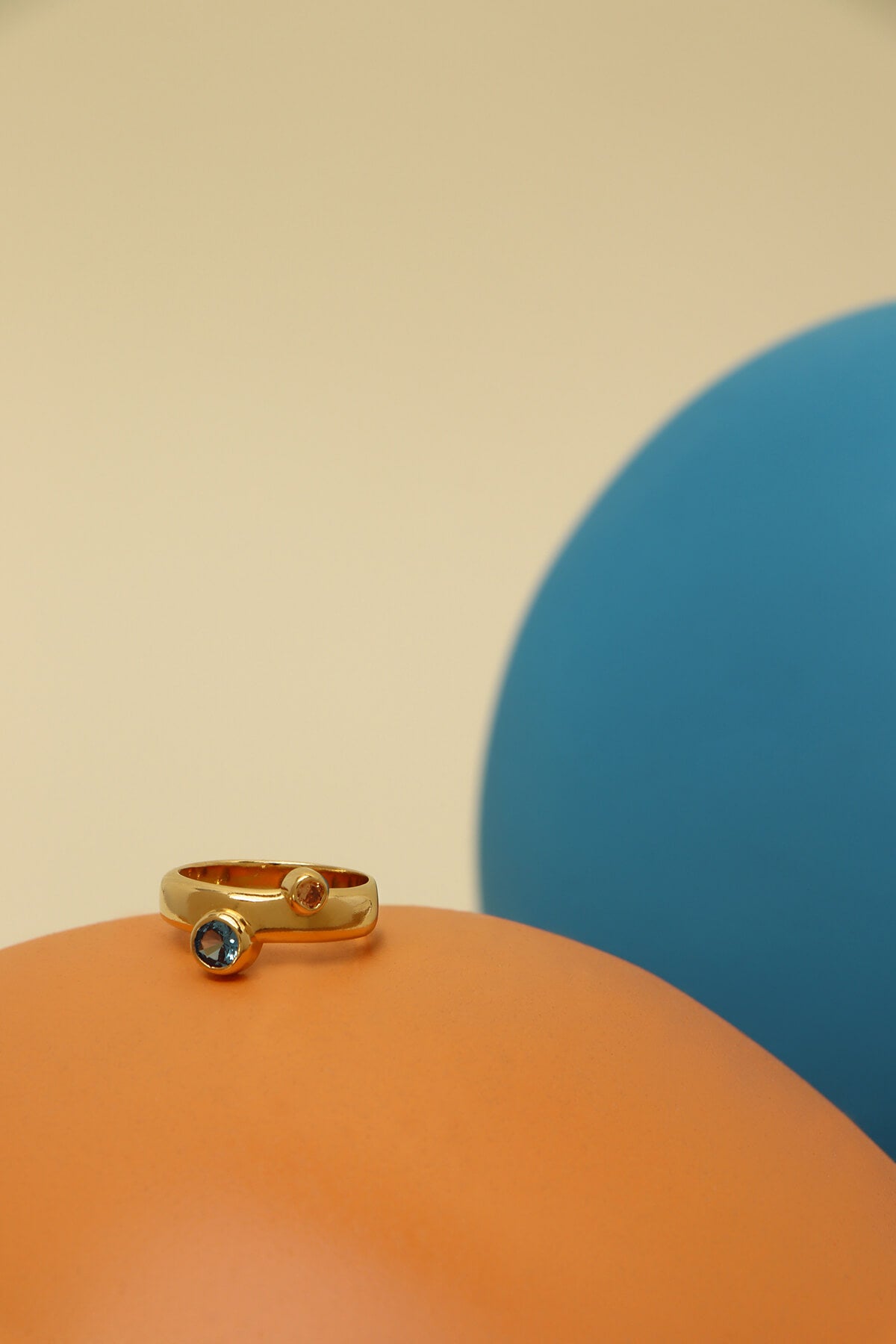 Bubble ring with cubic zirconia featuring blue and orange bezel-set stones on a polished band atop a smooth orange sculptural form.