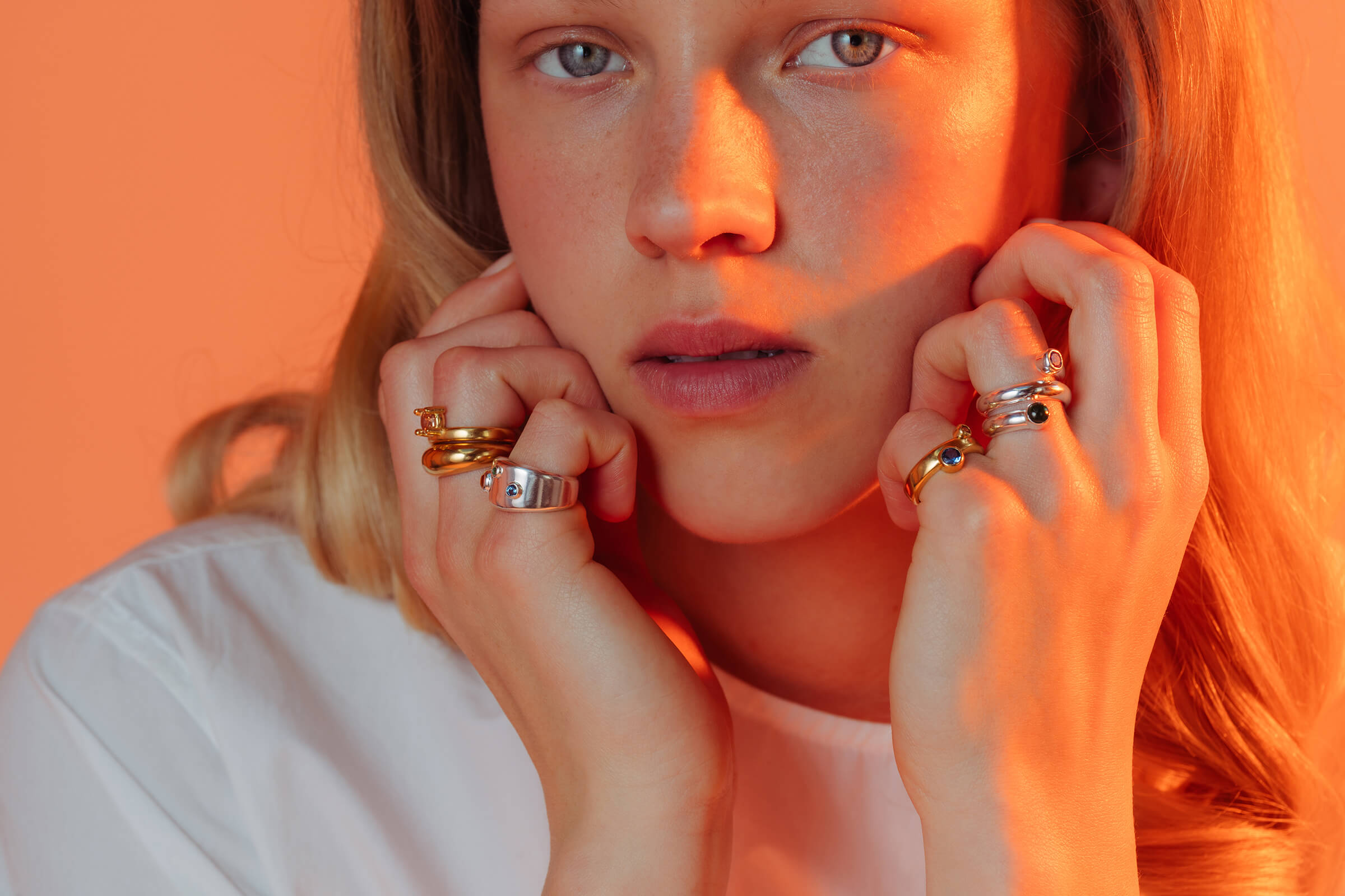 Drip drop rings worn stacked on both hands with coloured gemstones in a warm golden-hour portrait.