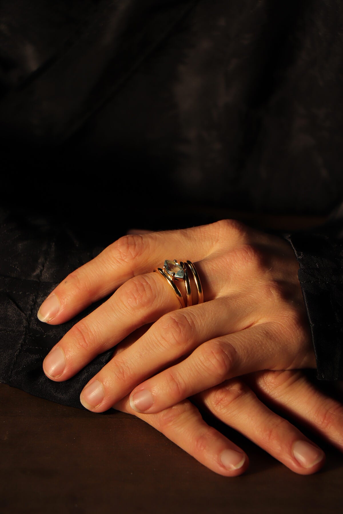 Twist ring with blue sky topaz, worn on the middle finger of a woman's hand. The ring features three slender, intertwining bands meeting at the faceted stone. Warm side lighting, dark background.